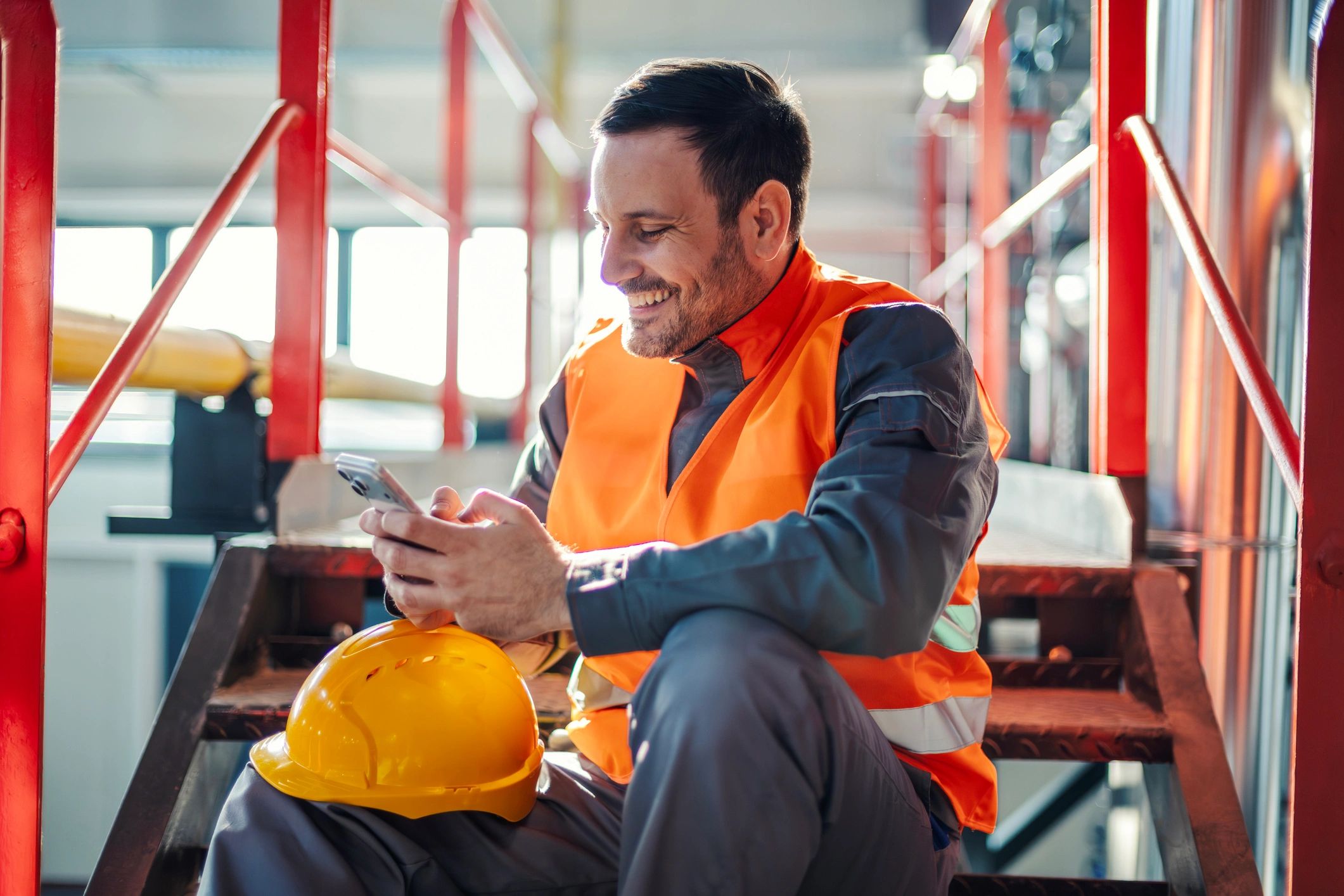 Smiling tradesperson wearing a safety vest and hard hat using a smartphone during a work break
